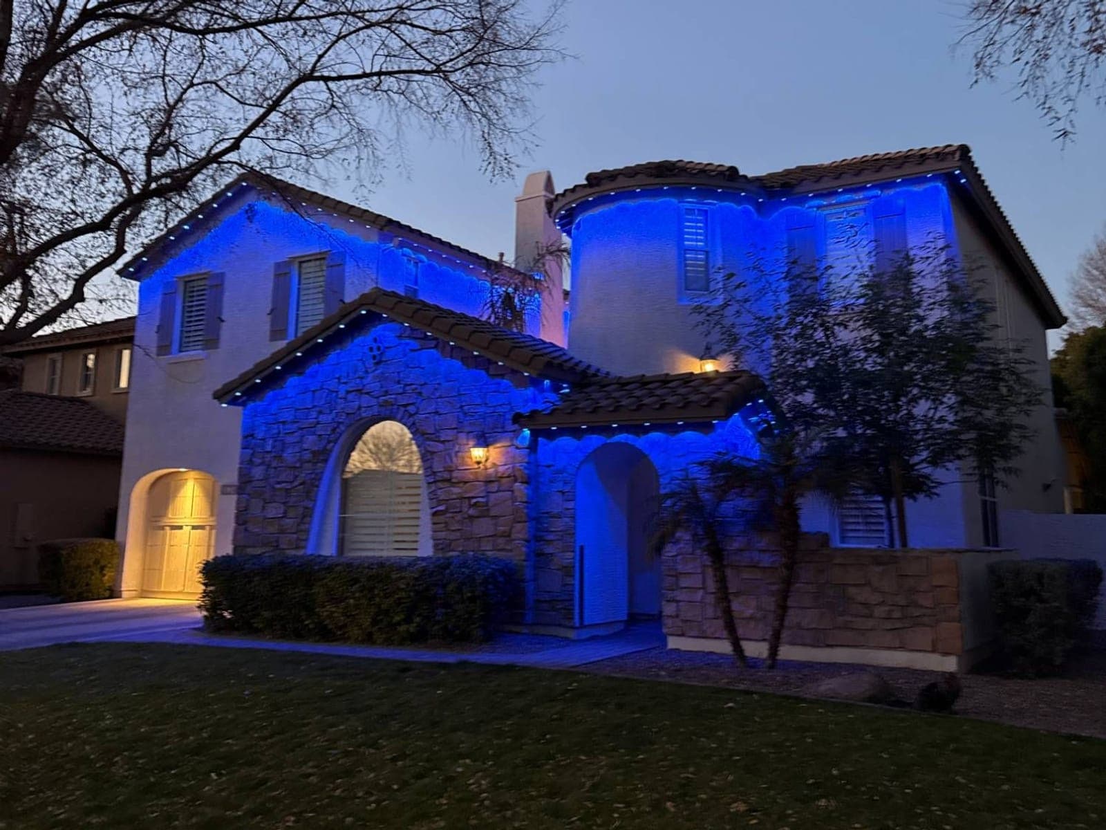 Two-story stucco and stone home with blue roofline lights at dusk