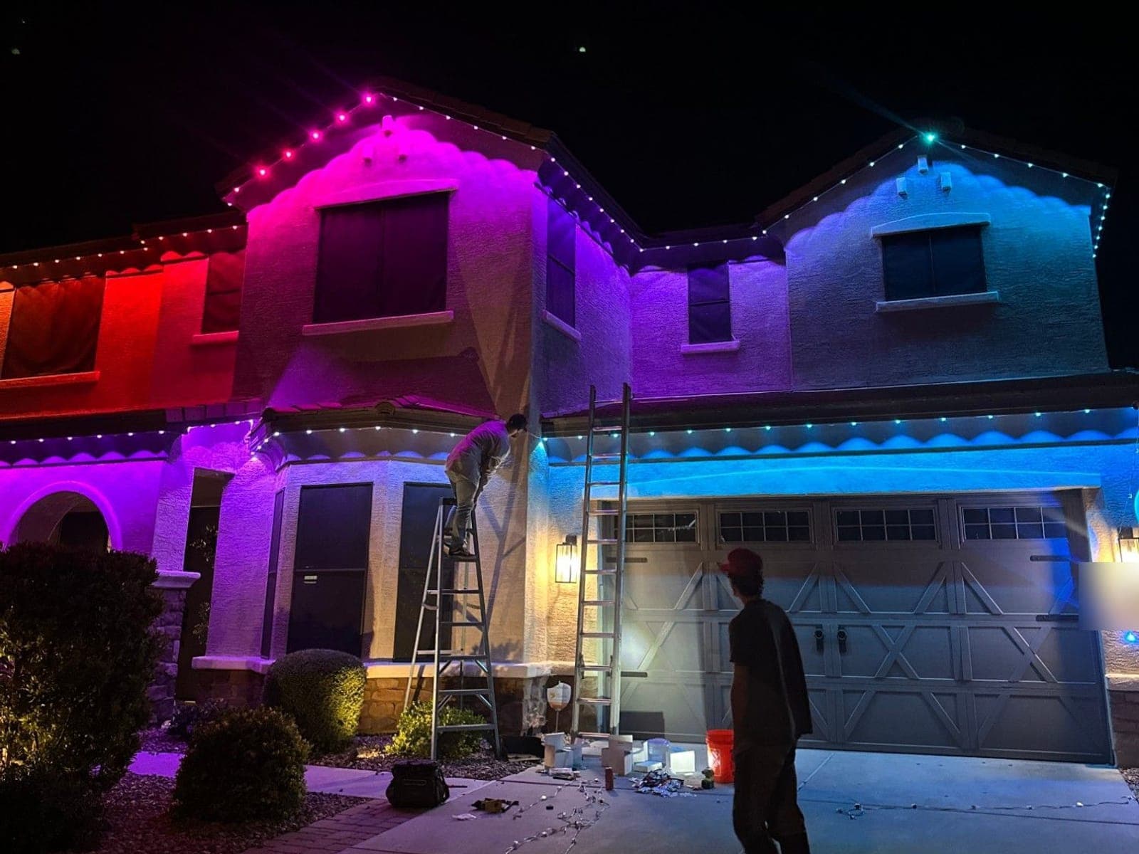 Crew installing multicolor roofline lights on a two-story home at night